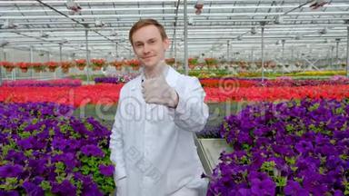 一位年轻的<strong>男科</strong>学家或农学家，穿着一件白色外套，在温室里放着花的床的背景下工作